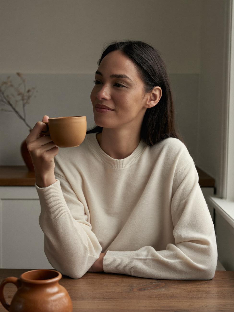 A woman holding a warm mug at her kitchen table in soft morning light