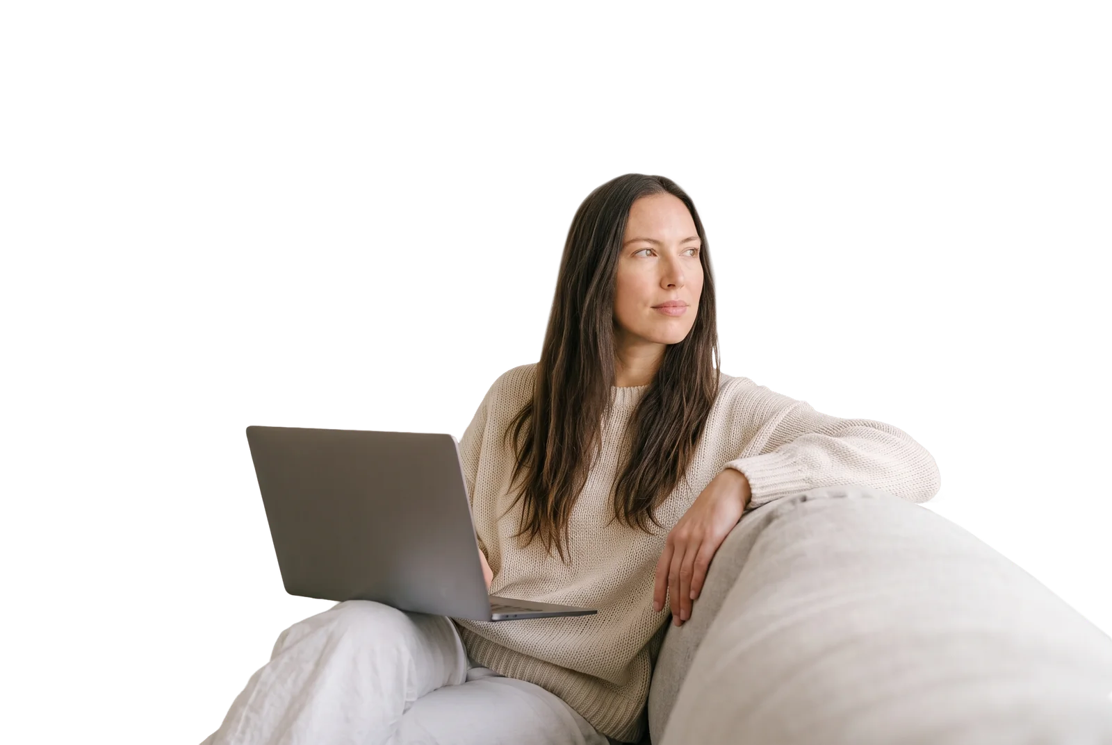 A woman in a cream knit sweater relaxes on a sofa with a laptop on her lap, looking thoughtfully into the distance